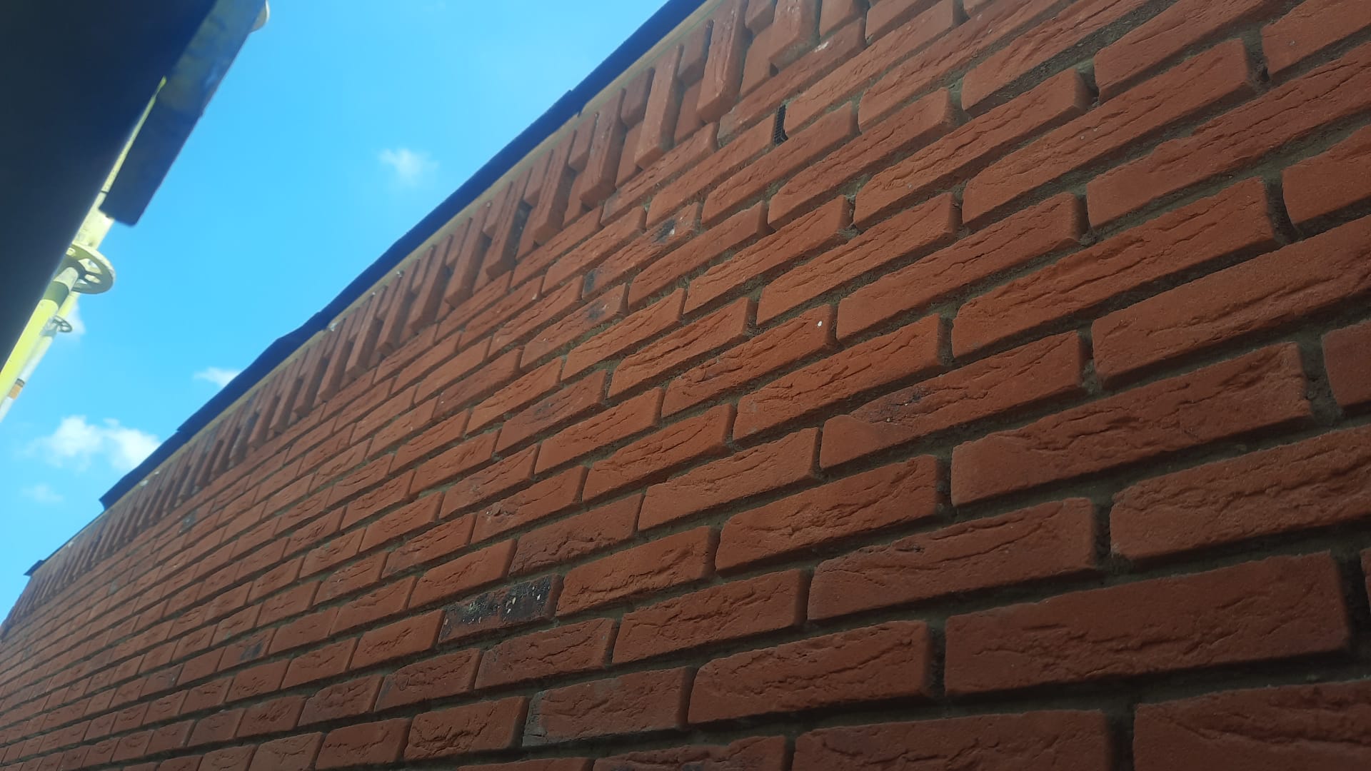 Rode bakstenen gevel met decoratief siermetselwerk onder de dakrand, tegen een blauwe lucht (foto 1 van 3)