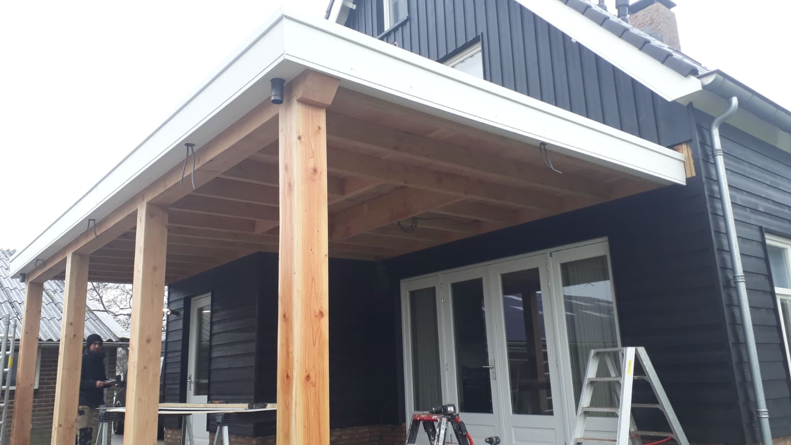 Houten overkapping met massieve staanders en balkenplafond aan een woning met zwarte gevelbekleding